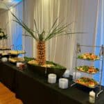 A buffet table with salads, appetizers, and a shrimp display arranged around a pineapple centerpiece, set against white curtains.
