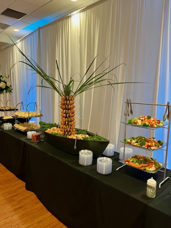 A buffet table with salads, appetizers, and a shrimp display arranged around a pineapple centerpiece, set against white curtains.
