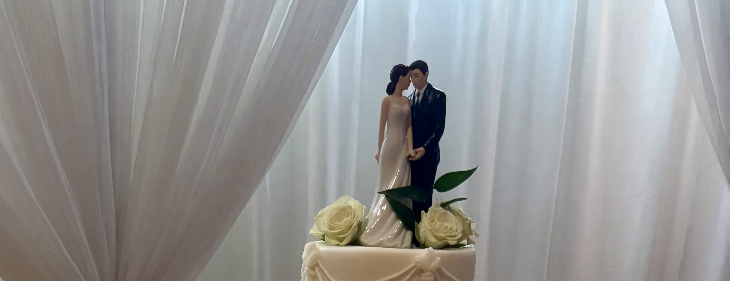 A wedding cake topper from Amy's Bridal featuring a bride and groom atop a cake adorned with white roses and draped icing.