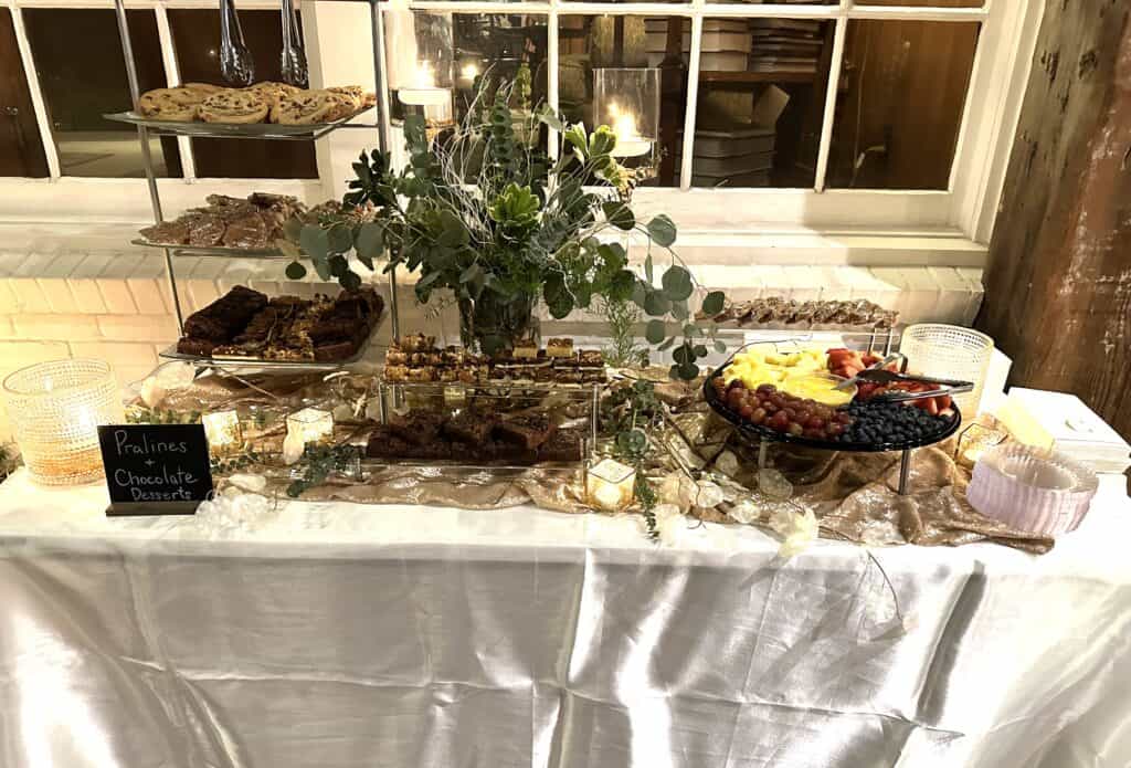 A dessert table with cookies, pralines, brownies, assorted desserts, and a fruit platter, decorated with greenery and candles.