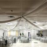 A banquet hall with white draped fabric on the ceiling, round tables with silver chairs, and minimal decorations.