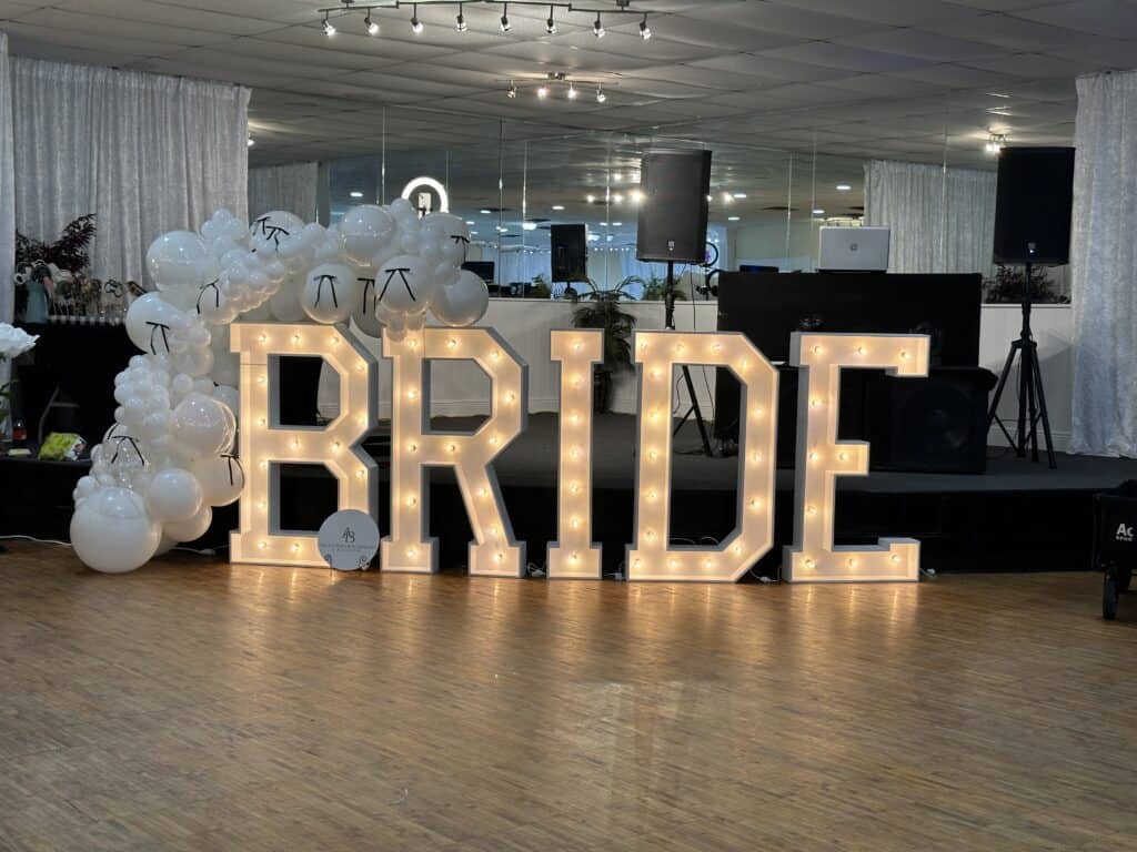 Large, illuminated letters spelling "BRIDE" are displayed indoors with white balloons and a DJ setup in the background.