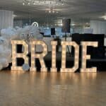 Large, illuminated letters spelling "BRIDE" are displayed indoors with white balloons and a DJ setup in the background.