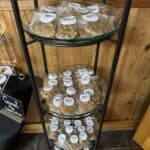 Three glass shelves on a metal rack display individually wrapped pralines with white labels in a wood-paneled room.