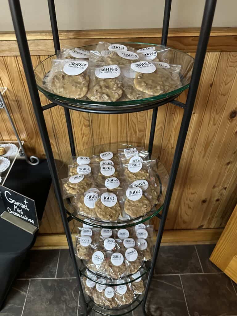 Three glass shelves on a metal rack display individually wrapped pralines with white labels in a wood-paneled room.