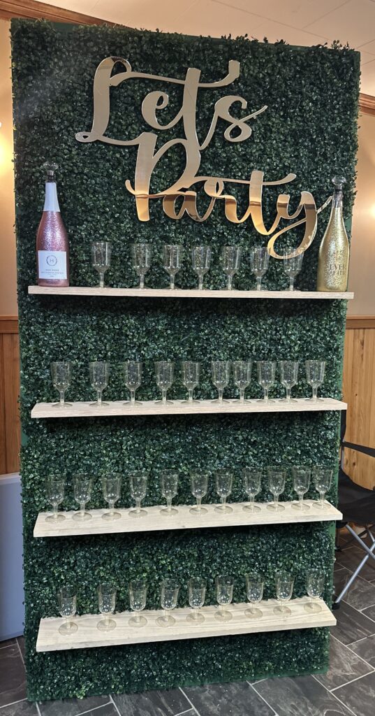 A green hedge wall with "Let's Party" sign, holding three shelves of empty champagne glasses and two bottles on top shelf.