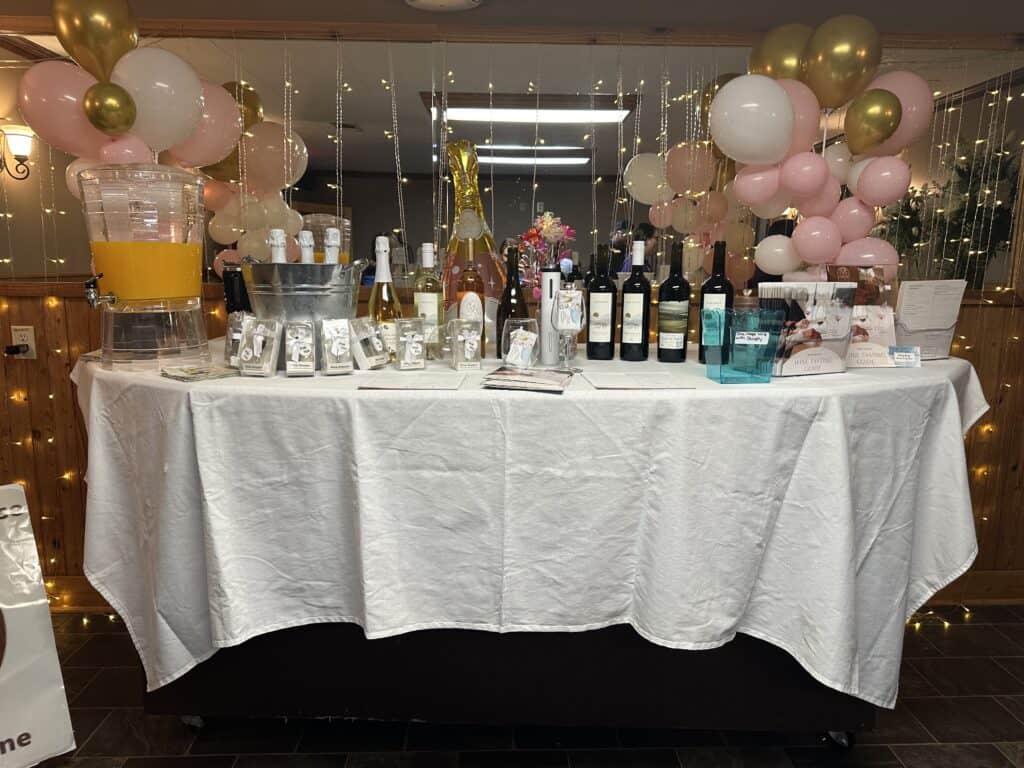 A table with a white tablecloth displays assorted wine bottles, drinks, cups, and balloons in pink, gold, and white decorations behind it.