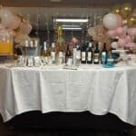 A table with a white tablecloth displays assorted wine bottles, drinks, cups, and balloons in pink, gold, and white decorations behind it.
