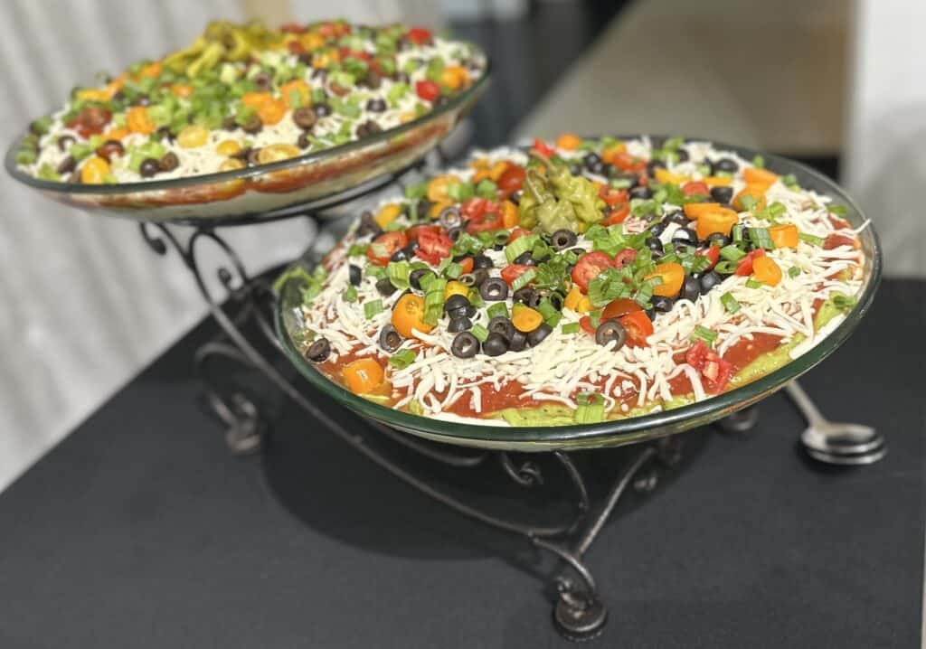 Two glass bowls of layered dip topped with shredded cheese, black olives, tomatoes, green onions, and guacamole on a black table.