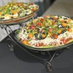 Two glass bowls of layered dip topped with shredded cheese, black olives, tomatoes, green onions, and guacamole on a black table.